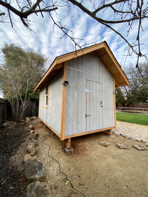 Backyard shed with cedar-toned siding and overhanging roof, corner view